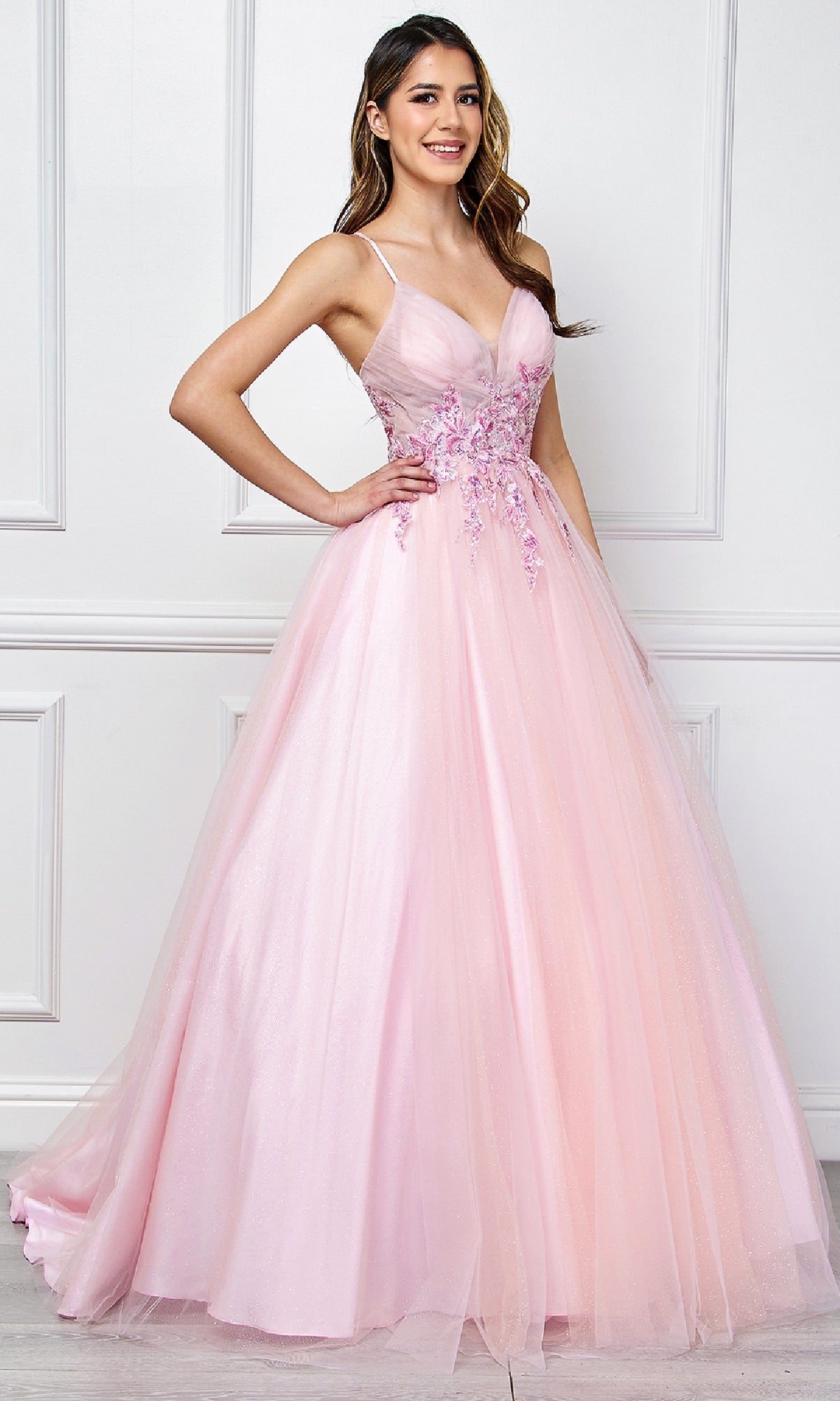 A woman wears the Amelia Couture SU088 Long Pastel Prom Ball Gown, featuring floral embroidery and a tulle skirt, standing against a white paneled wall with a smile and one hand on her hip.